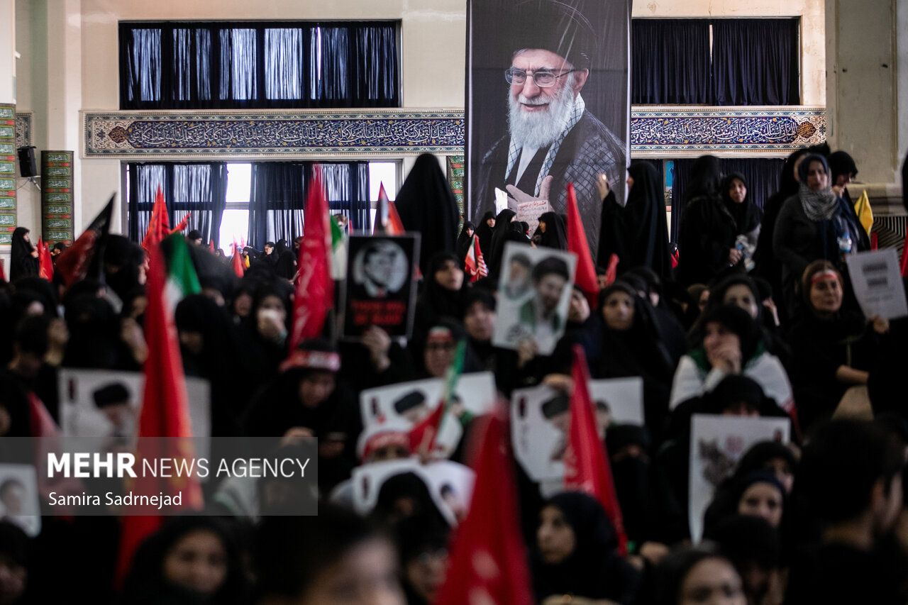 Mourners in black hold portraits and flags beneath a large photo of Ali Khamenei at a Tehran commemoration for the martyred Leader.