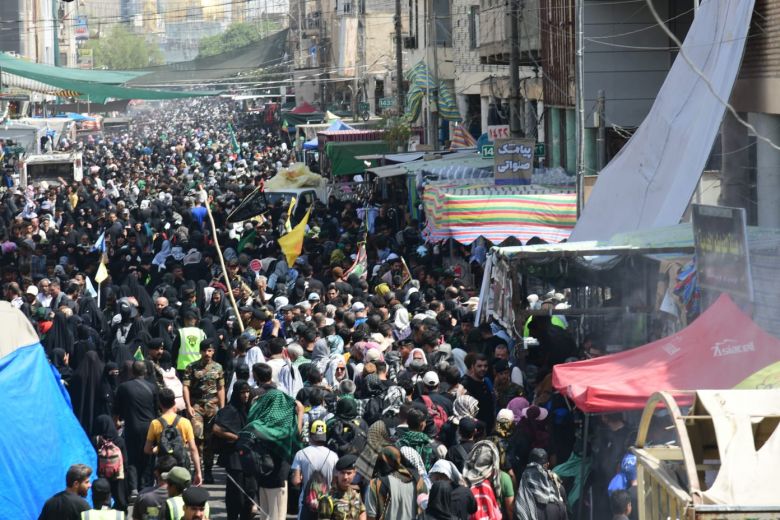 Crowds of black-clad pilgrims on the Arbaeen pilgrimage walk past roadside mawkib stalls and banners on a Karbala street in 2025.